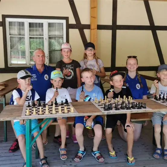 Eine Gruppe von Kindern und einem Erwachsenen spielt Schach zusammen auf einem Holztisch. Die Kinder sitzen auf dem Boden, tragen Mützen und Turnschuhe. Hinter ihnen steht eine Frau und lächelt. Die Umgebung ist im Freien, mit einem Holzhaus und einem Baum im Hintergrund.