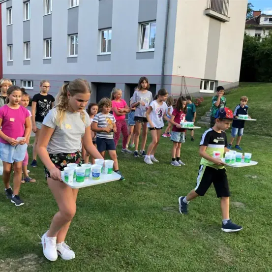 Kinder halten Tabletts mit Bechern vor einem Gebäude. Ein Mädchen und ein Junge laufen mit den Tabletts auf dem Rasen.