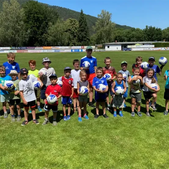 Eine Gruppe von Kindern, einige mit Mützen, stehen auf einem Rasenfeld und halten Fußballbälle, mit Bäumen und einem Gebäude im Hintergrund.