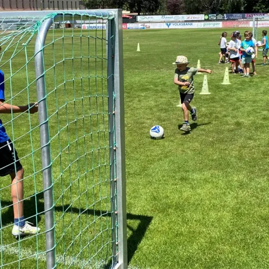Ein Junge tritt einen Fußball auf einem Feld, mit einem Torpfosten hinter ihm. Andere Kinder sind im Hintergrund und üben mit Kegeln.