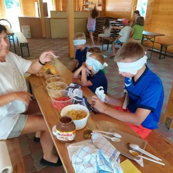 Mehrere Kinder haben Augenbinden auf und essen Essen aus Schüsseln auf einem Holztisch. Eine Frau hält einen Löffel. Ein Glas Nutella steht auf dem Tisch.