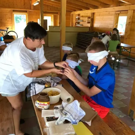 Eine Frau verblindet einen Jungen am Tisch mit Essen, andere Kinder und Erwachsene sind in einem Holzraum anwesend.