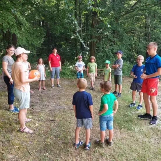 Eine Gruppe von Erwachsenen und Kindern versammelt sich in einer Lichtung in einem bewaldeten Gebiet. Eine erwachsene Frau hält einen Ball und schaut den Kindern ins Gesicht. Die Kinder schauen aufmerksam zu.