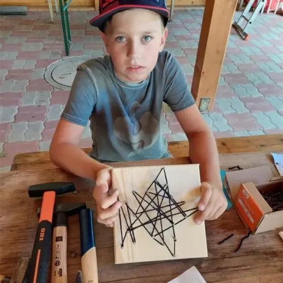 Ein junger Junge sitzt an einem Holztisch und hält eine Holzplatte mit einem geometrischen Muster. Er trägt eine Baseballkappe und ein graues T-Shirt. Neben ihm befinden sich mehrere Hämmer, Nägel und eine Schachtel.