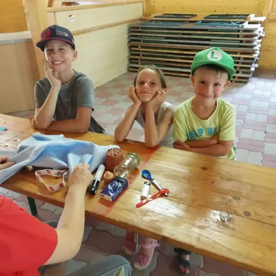 Drei Kinder sitzen mit einer Frau an einem Tisch. Einer hält einen Marker, ein anderer hat ein Maßband und der dritte ein blaues Tuch. Sie lächeln.