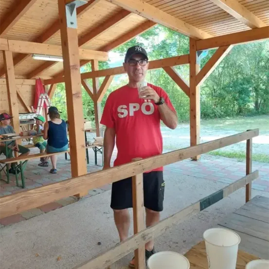 Ein Mann in einem roten T-Shirt und schwarzen Shorts steht in einem hölzernen Pavillon, hält einen Becher und hat andere Personen am Tisch im Hintergrund.
