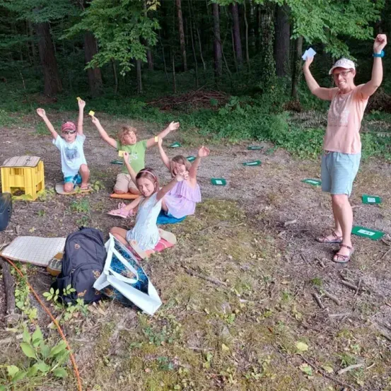 Ein Erwachsener und fünf Kinder sind im Freien in einem Waldgebiet und beschäftigen sich mit einer Aktivität. Der Erwachsene steht mit erhobenen Armen, während die Kinder auf dem Boden sitzen, einige mit erhobenen Händen. Sie sind von grünen Karten, Rucksäcken und einem Holzkisten umgeben.