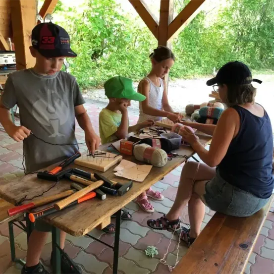 Vier Personen, darunter eine Frau und drei Kinder, arbeiten an einem Holztisch im Freien. Sie haben Werkzeuge und Garn auf dem Tisch. Die Kinder tragen Mützen und Schuhe. Die Frau trägt eine Brille und ein ärmelloses Oberteil. Sie sind wahrscheinlich mit einer Bastelaktivität beschäftigt.