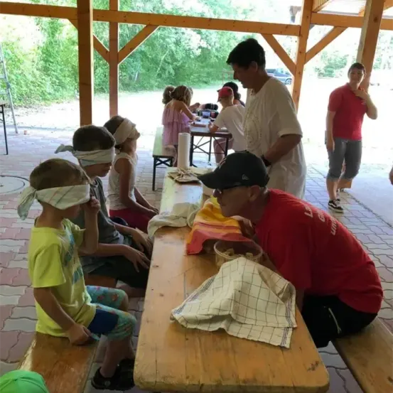 Geblendete Kinder sitzen auf Bänken in einem Pavillon, eines isst. Erwachsene stehen um einen Tisch. Im Hintergrund sitzt ein Mädchen an einem Tisch und ein Mann lehnt sich gegen eine Säule. Bäume und ein Auto sind draußen sichtbar.