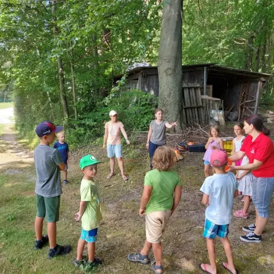 Eine Gruppe von Kindern und Erwachsenen steht in einem Grasbereich mit einem Schuppen im Hintergrund. Einige Kinder tragen Hüte, während andere barfuß sind. Eine erwachsene Frau in Rot spricht mit der Gruppe.