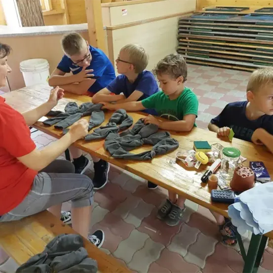 Fünf Jungen sitzen um einen Tisch herum, möglicherweise in einer Werkstatt. Ein Junge sieht sich gefaltete Kleidung an, während die anderen zuhören.