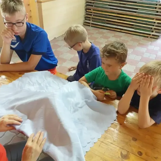 Fünf Jungen sitzen um einen Holztisch herum und untersuchen einen großen weißen Stoff mit gezackten Kanten. Zwei von ihnen tragen eine Brille.