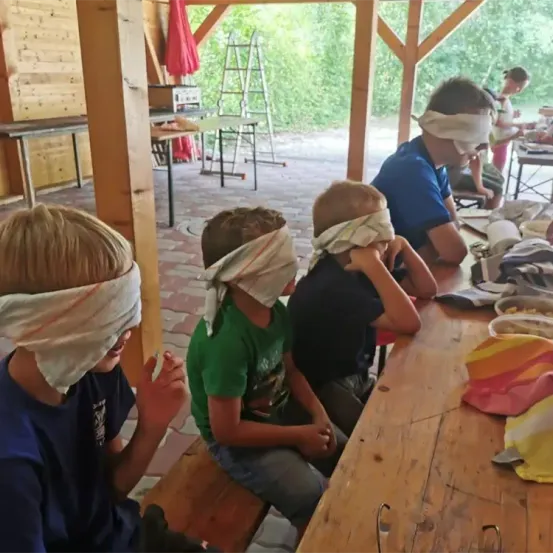 Vier Kinder mit verbundenen Augen sitzen um einen Tisch. Ein Kind hat die Hände im Gesicht. Ein Leiter und ein Tisch mit Gegenständen stehen hinter ihnen.