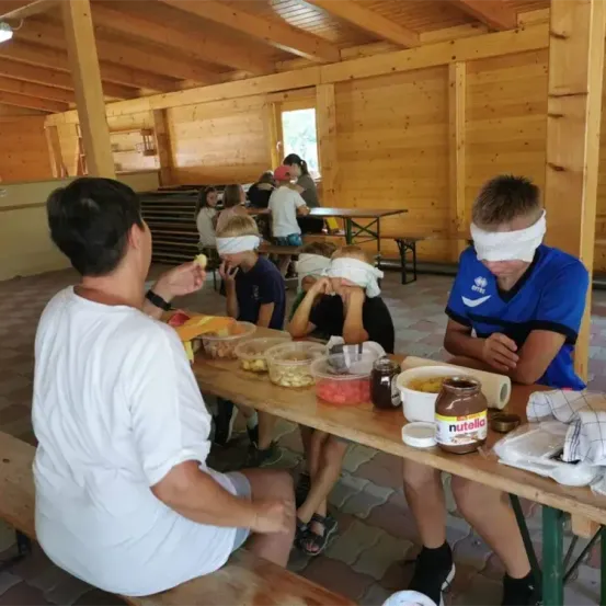 Mehrere Menschen mit verbundenen Augen sitzen an einem Tisch in einem hölzernen Raum. Sie essen und es gibt eine Nutella-Glas auf dem Tisch.