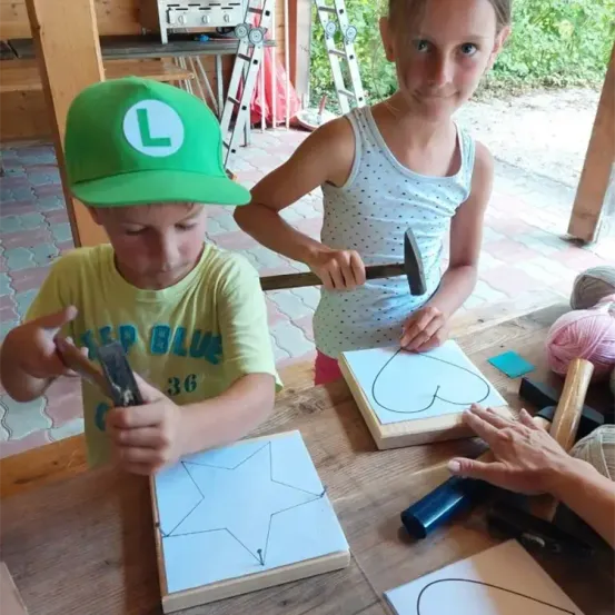 Zwei Kinder arbeiten mit Hämmern an Holzbrettern, eines hämmert ein Sterndesign, während das andere an einem Herz arbeitet. Beide tragen Freizeitkleidung und scheinen konzentriert zu sein.