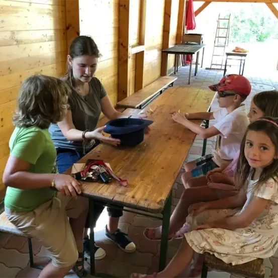 Eine Frau steht an einem Tisch mit mehreren Kindern um sie herum. Sie sitzen auf Bänken. Die Frau hält einen blauen Hut und scheint den Kindern etwas zu erklären.