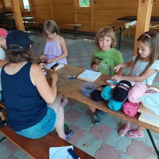 Eine Gruppe von Kindern arbeitet mit Hilfe eines Erwachsenen an einem Bastelprojekt, das um einen Holztisch herum sitzt. Verschiedene Bastelmaterialien liegen auf dem Tisch, und die Kinder benutzen Werkzeuge. Die Umgebung ist ein hölzerne Pavillon im Freien.