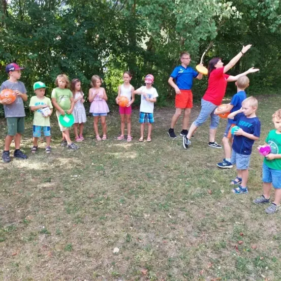 Eine Gruppe von Kindern und Erwachsenen spielt mit Bällen auf einer Rasenfläche mit Bäumen im Hintergrund.