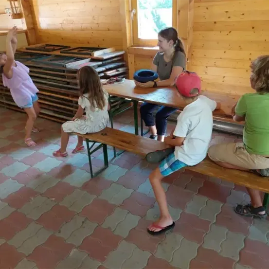 In einem hölzernen Raum sitzen Kinder auf Bänken. Ein Mädchen in einem lila Shirt steht hinter ihnen. Eine Frau sitzt an einem Tisch und hält eine blaue Schüssel. Holzkisten sind dahinter gestapelt.