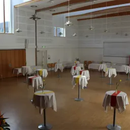 Ein leerer Saal mit Holzboden, weißen Wänden und mehreren Tischen mit weißen und roten Tischdecken, jeweils mit einer gelben Blume geschmückt. Es gibt auch Stühle und eine Tafel an der Wand.