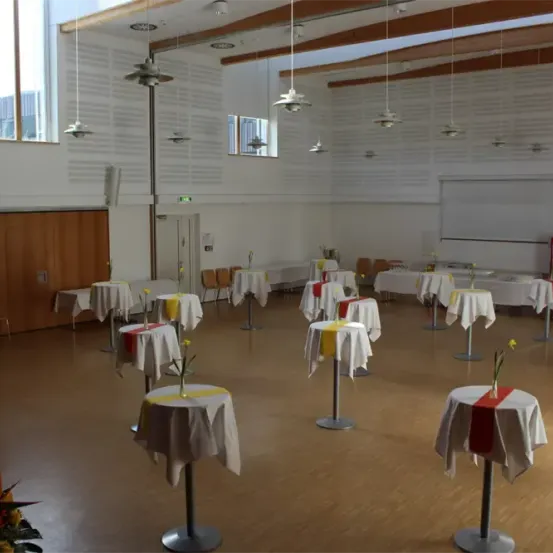 Ein leerer Saal mit Holzboden, weißen Wänden und mehreren Tischen mit weißen und roten Tischdecken, jeweils mit einer gelben Blume geschmückt. Es gibt auch Stühle und eine Tafel an der Wand.