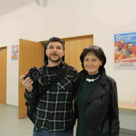 Ein Mann und eine Frau stehen in einem Raum und lächeln. Der Mann hält eine Kamera. Hinter ihnen ist ein Plakat. Türen und ein Tisch sind im Hintergrund.