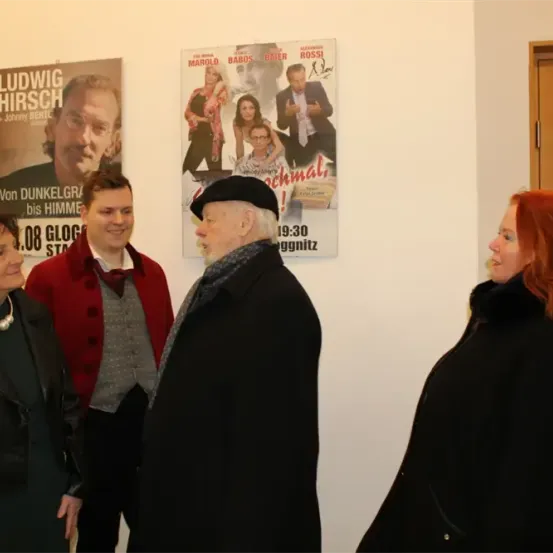 Vier Personen stehen vor einer Wand mit Filmplakaten. Der Mann in der Mitte trägt einen schwarzen Hut. Die Frau auf der rechten Seite hat rote Haare.