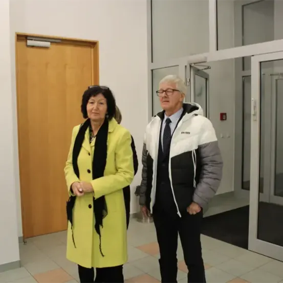 Ein älterer Mann und eine ältere Frau stehen in einem Flur mit weißen Wänden und gefliesten Böden. Die Frau trägt einen gelben Mantel mit einem schwarzen Schal, und der Mann trägt einen weißen und grauen Mantel. Hinter ihnen befinden sich zwei Türen, von denen eine offen ist.