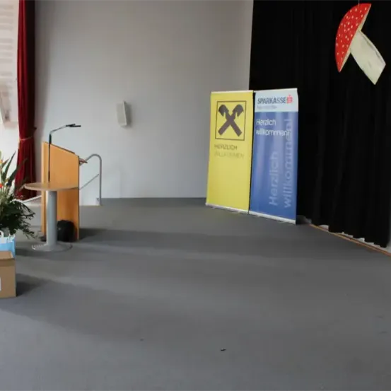 Ein leerer Raum mit einem Podium, Mikrofon und Bannern. Eine Blumenarrangement und eine Box sind links. Die Wand hat einen Lautsprecher und einen Pilz.