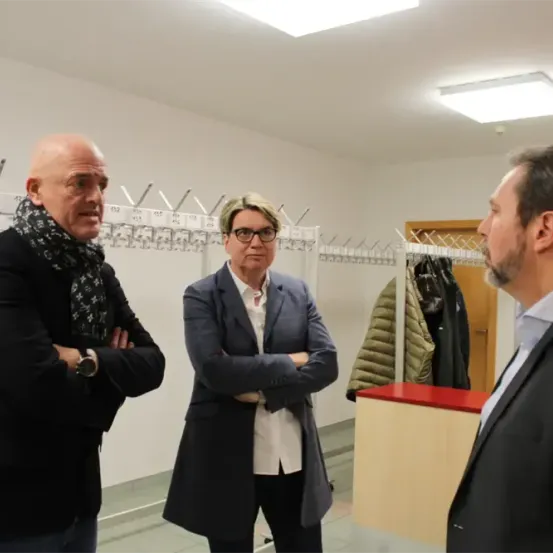 Drei Männer in formeller Kleidung stehen in einem Flur mit einem Metallkleiderständer und hängenden Jacken. Der Mann auf der linken Seite trägt einen Schal, die Frau in der Mitte trägt eine Brille und der Mann auf der rechten Seite schaut sie an.