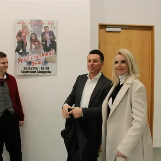 Drei Personen stehen vor einer Wand mit einem Plakat einer Theateraufführung, datiert auf den 20.4.2016. Ein Mann in einem roten Mantel, ein Mann in einem schwarzen Anzug und eine Frau in einem weißen Mantel.