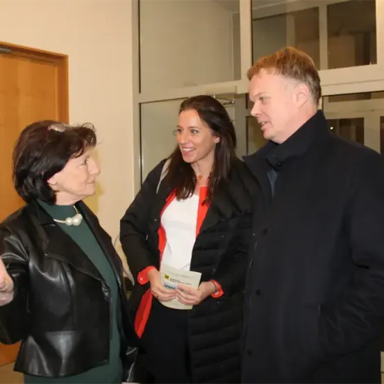 Drei Menschen stehen drinnen und unterhalten sich. Eine Frau gestikuliert mit der Hand. Eine andere Frau hält einen Becher. Der Mann schaut die Frau rechts von ihm an.