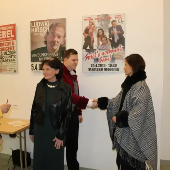Zwei Frauen schütteln sich die Hände in einem Raum mit mehreren Postern an der Wand. Ein Poster zeigt Ludwig Hirsch, ein anderes ein Bild eines Mannes und einer Frau mit Text darüber. Ein Tisch mit einem Stift und Papier ist im Vordergrund.