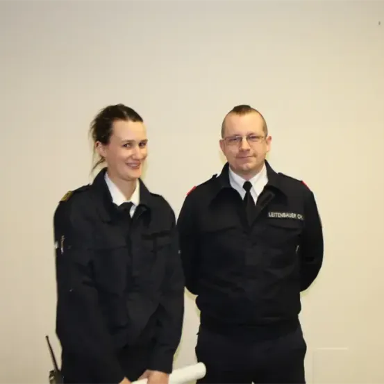 Zwei Personen in schwarzen Uniformen, wahrscheinlich Polizeibeamte, stehen eng beieinander und lächeln. Sie halten ein gerolltes weißes Objekt. Eine Frau trägt eine Abzeichen an ihrer Schulter. Ein Mann trägt einen Namensschild. Sie stehen vor einer weißen Wand.