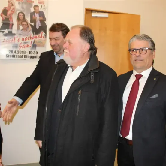 Drei Männer in Anzügen stehen gemeinsam vor einem Plakat, das für eine Show namens "Aniel's nachmals Sam!" wirbt. Das Datum und die Uhrzeit der Show sind auf dem Plakat angegeben.