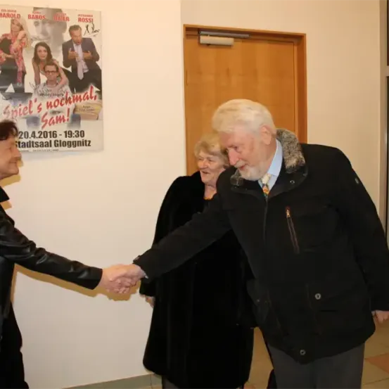 Ein älterer Mann mit weißen Haaren schüttelt die Hand einer Frau in einer schwarzen Lederjacke. Hinter ihnen ist ein Plakat, das einen Film bewirbt.