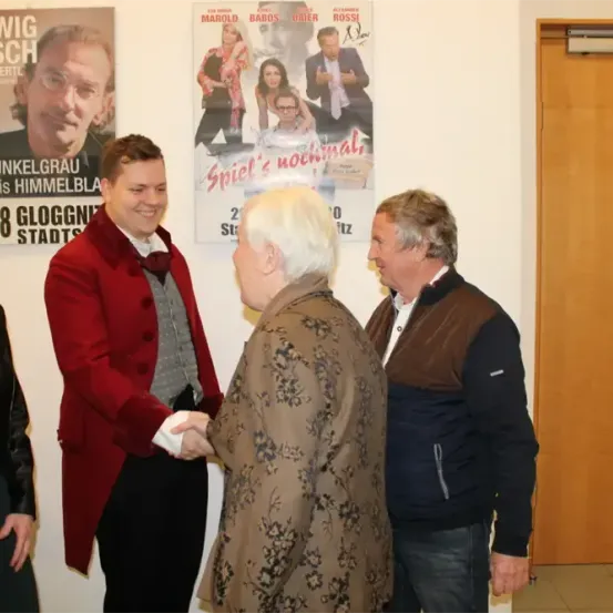 Vier Personen stehen vor einem Plakat, zwei von ihnen schütteln Hände. Einer trägt einen roten Mantel, eine andere ein Blumenkleid. Hinter ihnen zeigt ein Plakat einen Mann mit Brille.