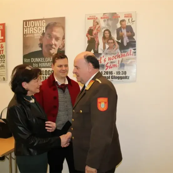 Eine Frau und ein Mann in einem roten Mantel schütteln sich die Hände mit einem Mann in Uniform, vor einer Wand mit Filmplakaten stehend.