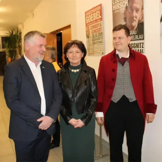 Drei Personen stehen in einem Flur mit dekorativen Postern an der Wand, eine in einer schwarzen Lederjacke, eine in einem roten Mantel und eine in einem Anzug.