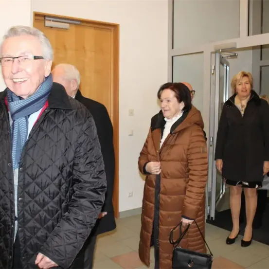 Eine Gruppe von Menschen, darunter ein älterer Mann und eine ältere Frau, steht lächelnd vor einer Tür. Die Frau trägt eine schwarze Handtasche.