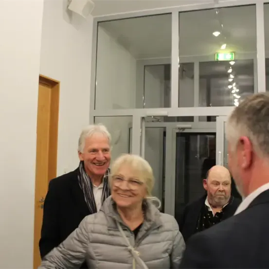 Drei Menschen stehen vor einer Glastür. Eine Frau lächelt und trägt eine Brille. Der Mann neben ihr trägt einen Schal. Ein anderer Mann schaut sie an. Hinter ihnen ein Poster mit einem Mann und dem Wort 'Bossi'.