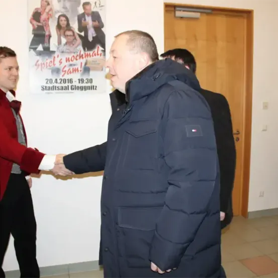 Zwei Männer schütteln sich die Hände in einem Raum mit einer weißen Wand, einem Poster und einer Holztür. Ein dritter Mann steht hinter ihnen.