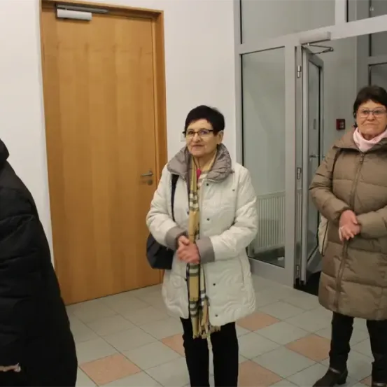 Drei Frauen stehen in einem Flur, eine mit Brille und Schal, eine andere in einem braunen Mantel. Hinter ihnen ist eine Tür und eine offene Glastür zu sehen.