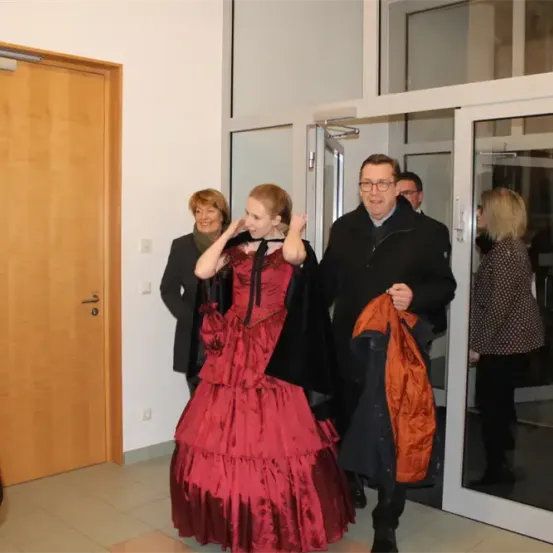 Eine Frau in einem roten Kleid steht neben einer Tür, während ein Mann in einem schwarzen Mantel einen roten Mantel hält. Hinter ihnen lächelt eine Frau in die Kamera.