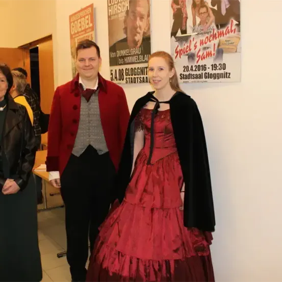 Drei Personen stehen zusammen vor einer Wand mit verschiedenen Postern, darunter eines mit einem Mann und ein anderes mit dem Text Spiel's noch mal, Sam! und einem Datum.