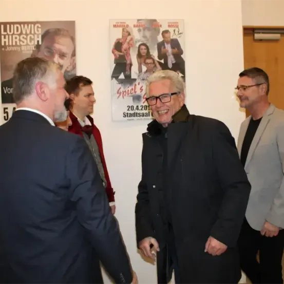 Vier Männer stehen vor einer Wand mit zwei Postern. Ein Mann in einem schwarzen Mantel lächelt und sieht einem anderen Mann in einem Anzug entgegen.
