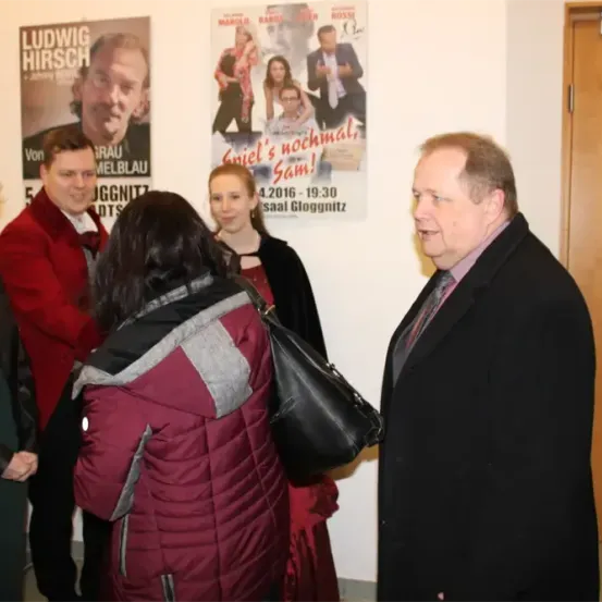 Eine Gruppe von Menschen steht vor einer Wand mit Filmplakaten. Der Mann rechts trägt einen schwarzen Anzug und eine Krawatte.