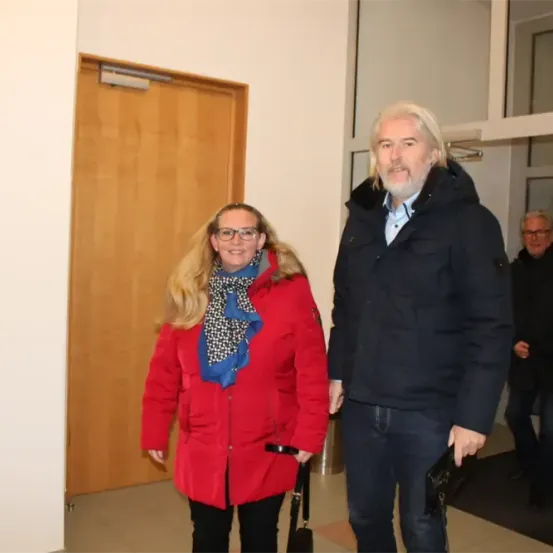 Ein Mann und eine Frau stehen vor einer Tür. Die Frau trägt einen roten Mantel und einen Schal. Der Mann ist in einem schwarzen Jackett und hat ein Lächeln im Gesicht. Hinter ihnen steht ein weiterer Mann.