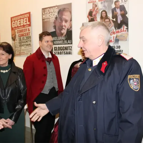 Drei Personen stehen in einem Raum mit Postern an der Wand. Einer in Polizeiuniform spricht mit einer Frau in einer schwarzen Lederjacke.