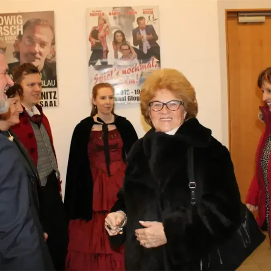 Eine Gruppe von Menschen, darunter eine Frau in einem schwarzen Pelzmantel, steht in einem Raum mit einer Tür und Postern im Hintergrund.